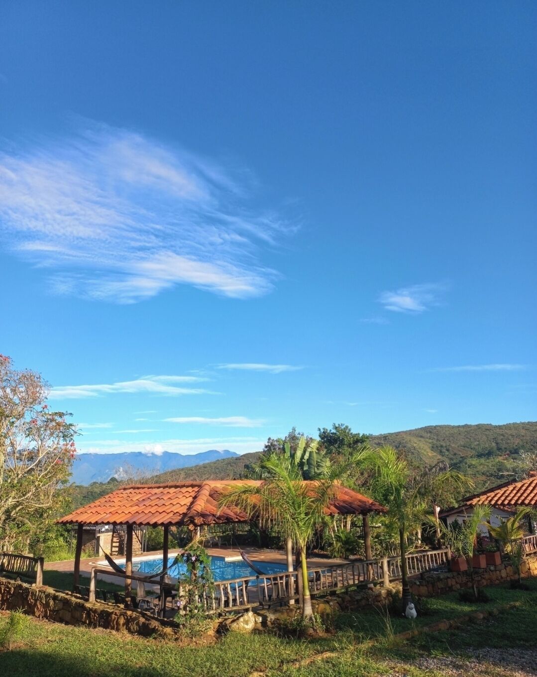 Zona de piscina con cubierta de madera y un área de estar techada, rodeada de vegetación exuberante y colinas al fondo.