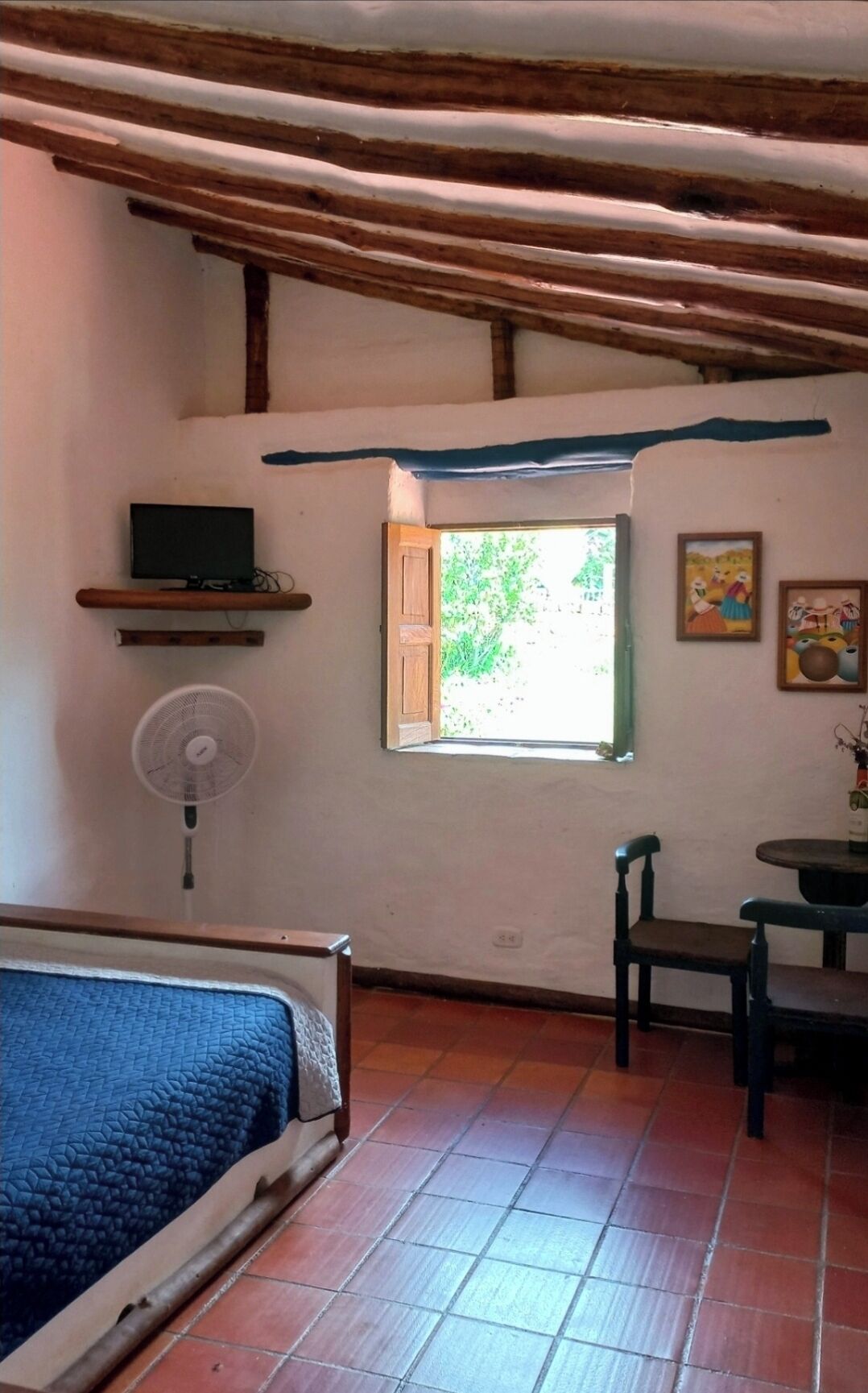 A room with a bed, a fan, a TV on a shelf, a window with a view of greenery, and a table with chairs.