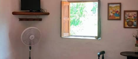 A room with a bed, a fan, a TV on a shelf, a window with a view of greenery, and a table with chairs.