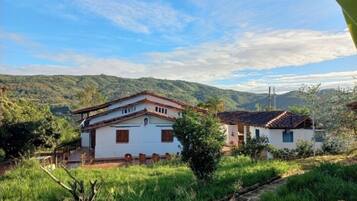 Una casa rural de paredes blancas y techo rojo, rodeada de exuberante vegetación y colinas al fondo.
