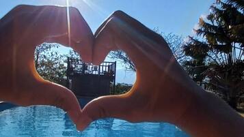 Una mano forma un corazón frente a una piscina, con el sol brillando al fondo.