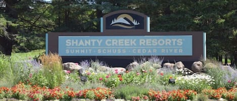 Placa para Shanty Creek Resorts com logotipo de montanha e rio, rodeada por um jardim paisagístico com diversas plantas e flores.