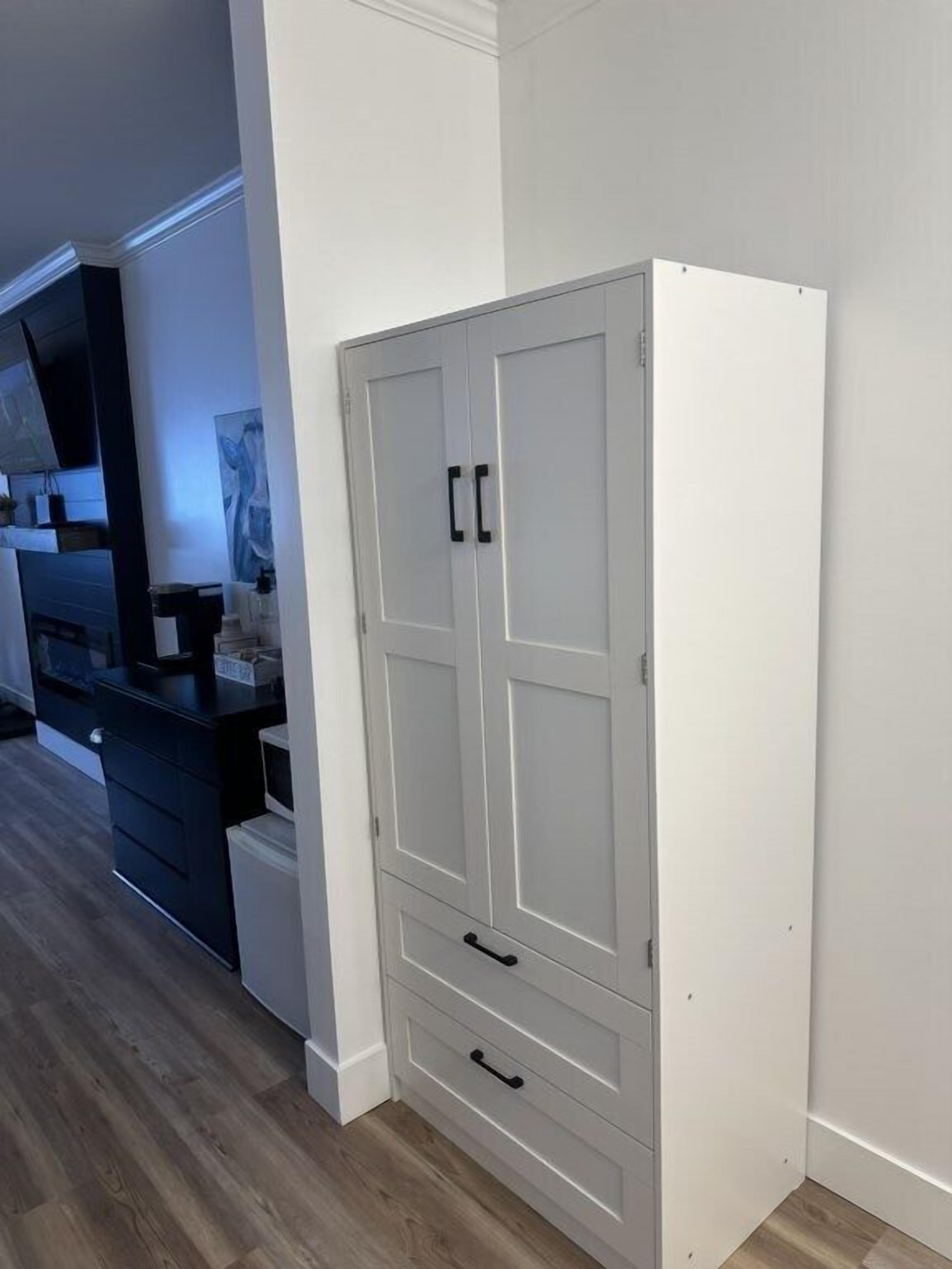 Um guarda-roupa branco com duas portas e duas gavetas, situado num quarto com soalho de madeira e uma cômoda escura à esquerda.
