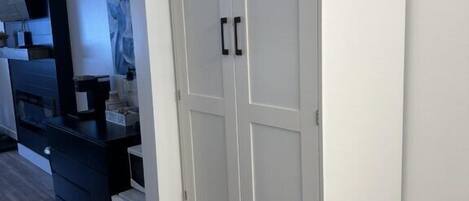 Um guarda-roupa branco com duas portas e duas gavetas, situado num quarto com soalho de madeira e uma cômoda escura à esquerda.