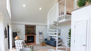 A spacious living room with a round dining table, grey upholstered chairs, a fireplace, a blue sofa, and a white spiral staircase.