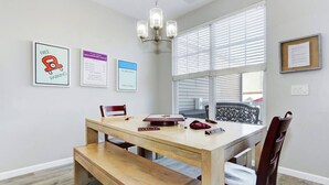 A dining area with a wooden table, benches, and chairs. There are framed signs on the wall, a chandelier, and a window with blinds.