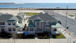 A row of three-story houses with green roofs and white trim, located near a beach and a road.