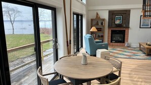 A spacious living room with a fireplace, a round wooden table, and a view of a lake through large windows. - Camp Wagoner Lake Cabins (Mayfield)
