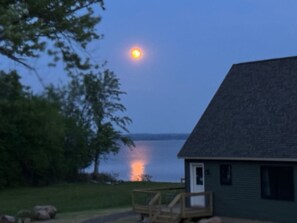 Une maison au bord d’un lac, au crépuscule, avec une lune visible.