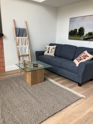Een moderne woonkamer met een glazen salontafel, een blauwe bank met decoratieve kussens, een houten ladder en een schilderij aan de muur.