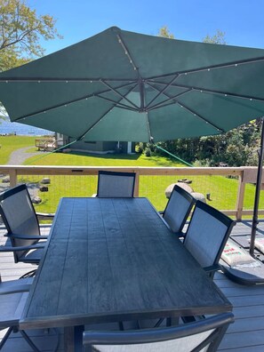 Una terraza de madera con una mesa y sillas, una sombrilla y vistas a una casa y vegetación.