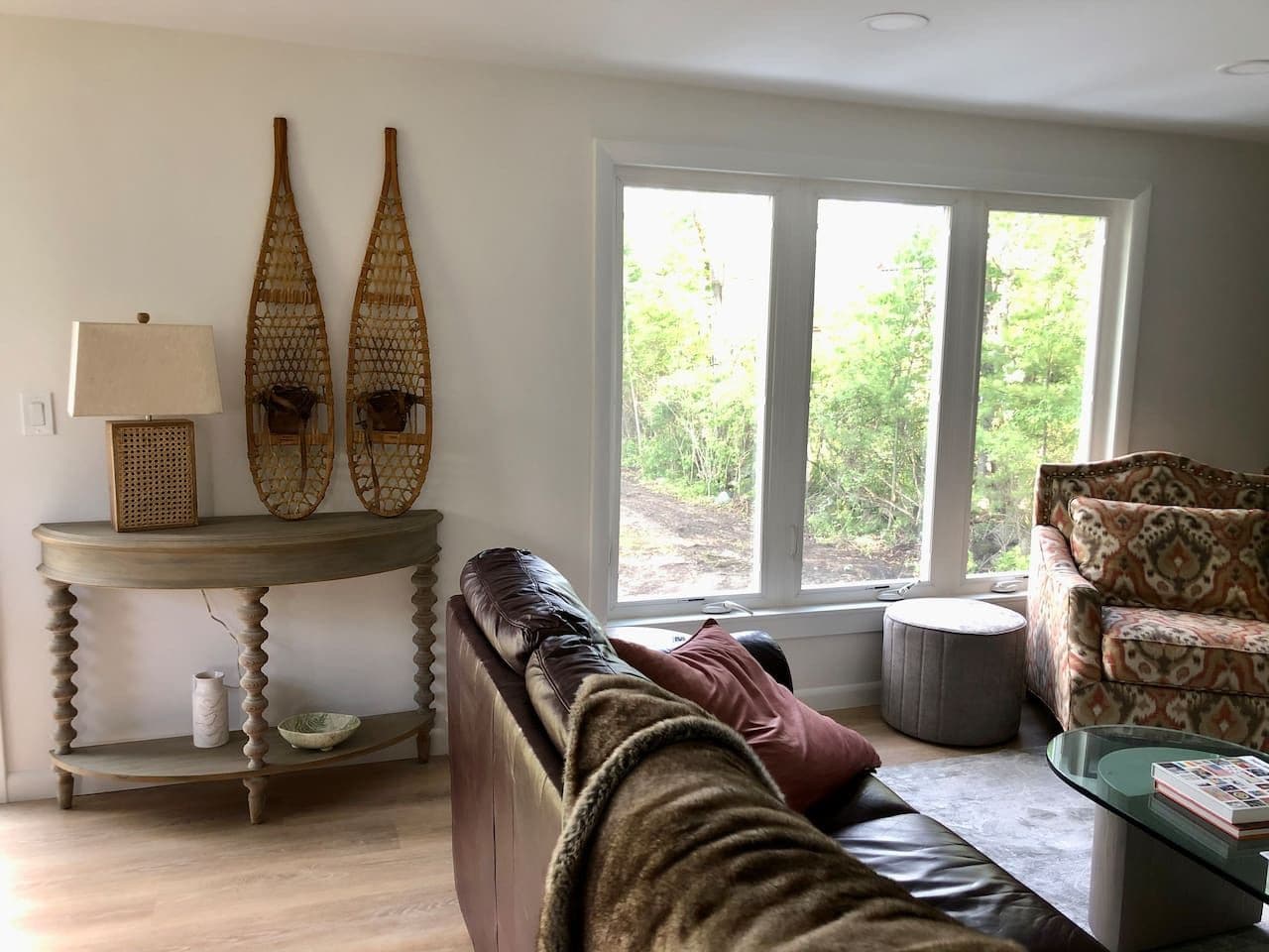 A cozy living room with a leather sofa, a round coffee table, and a patterned armchair. A window provides a view of greenery outside.