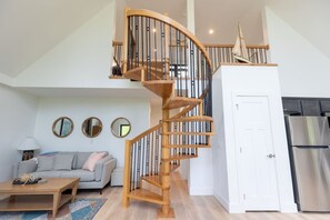 Uma sala de estar moderna com uma escada espiral de madeira, um sofá cinza, uma mesa de centro e uma cozinha com frigorífico de aço inoxidável.