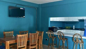 A room with a dining table and chairs, a TV mounted on the wall, a kitchenette with cabinets and a microwave, and a sofa.