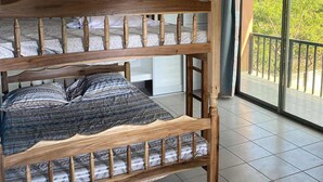 A bunk bed with a wooden frame and patterned bedding, a white wardrobe, and a balcony with a view of greenery.