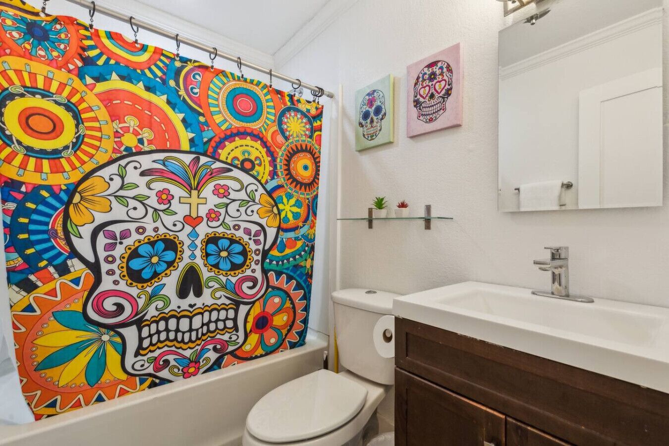 A bathroom with a colorful shower curtain, a white toilet, a sink with a mirror, and a small shelf with plants.