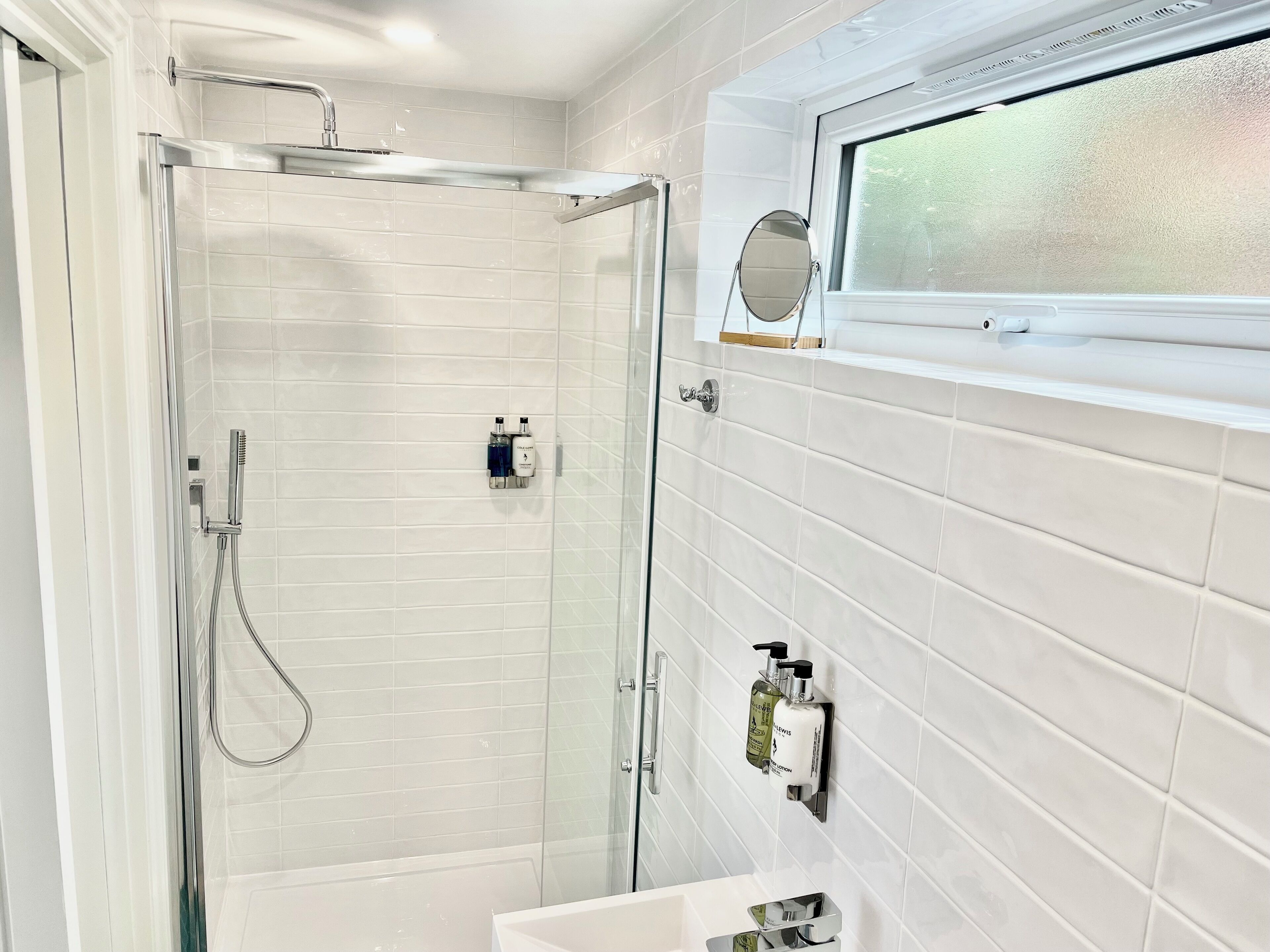A modern bathroom with a glass-enclosed shower, white tiled walls, and a window above the sink.