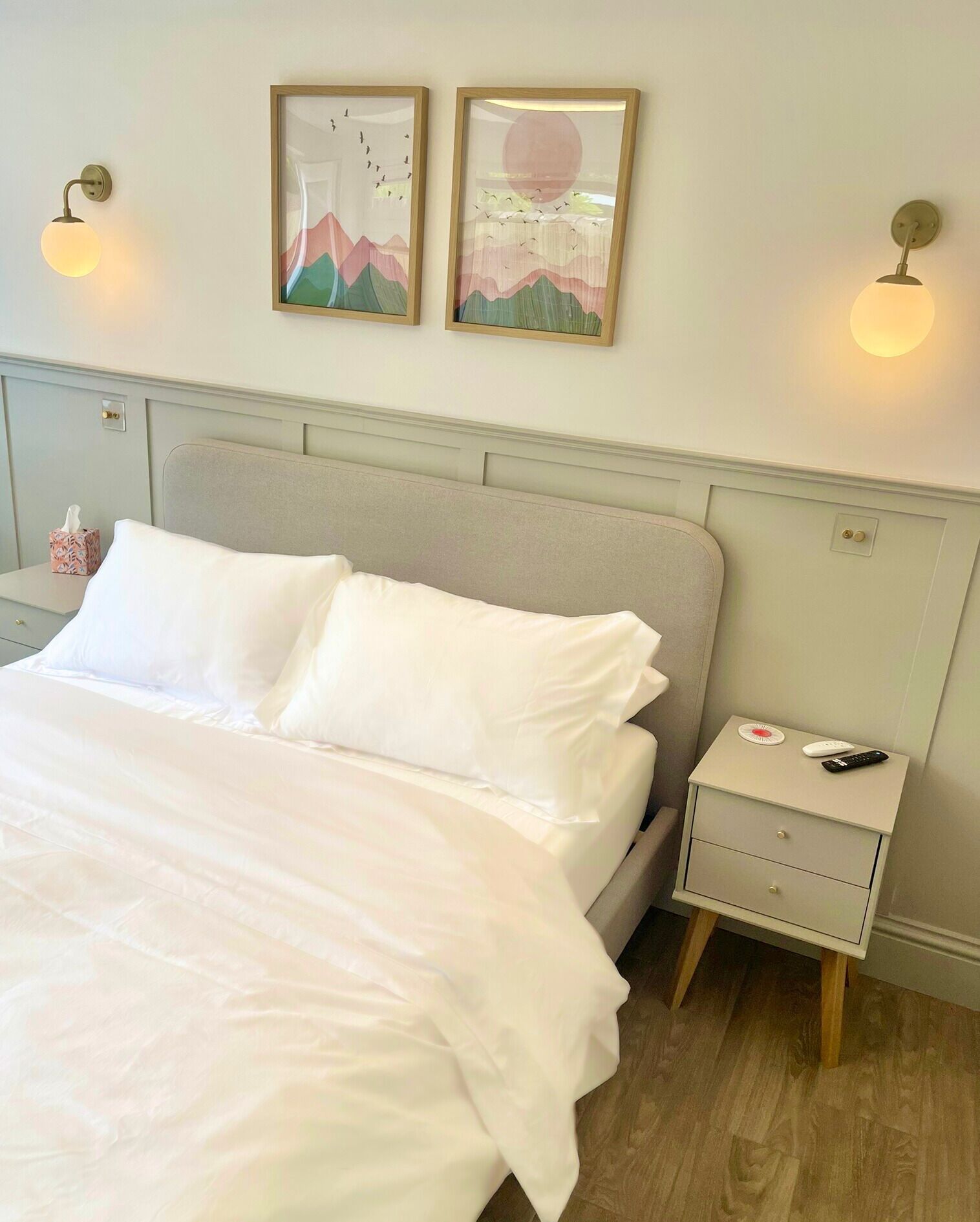 Een net opgemaakt bed met witte lakens in een kamer met een lichtgrijs hoofdeinde. Er zijn twee wandlampen en twee ingelijste kunstwerken boven het bed.