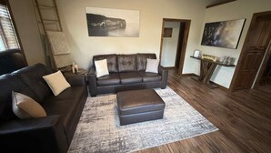 Ein Wohnzimmer mit einem dunkelbraunen Ledersofa, einem passenden Hocker und einem großen Teppich. Ein Leitgerüst lehnt an der Wand, über dem Sofa hängt ein Gemälde und eine Tür führt in einen anderen Raum.