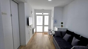 Una habitación moderna y luminosa con suelo de madera, un sofá oscuro, un escritorio pequeño y una televisión en la pared.