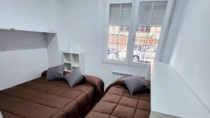 Una habitación pequeña y funcional con dos camas, una ventana con rejas, un radiador y un escritorio con silla.
