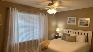 Una habitación de hotel con cama, mesita de noche con lámpara, ventilador de techo y ventana con cortinas.