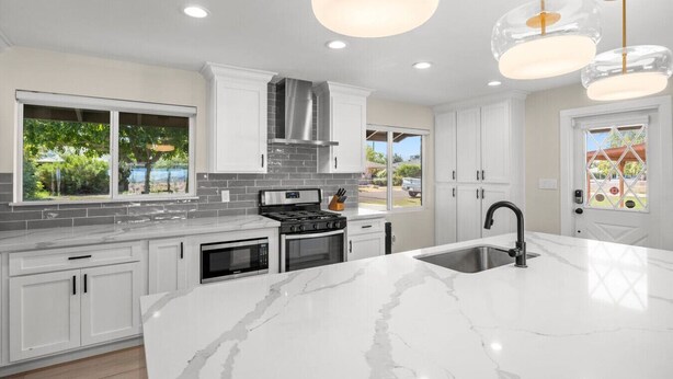 A modern kitchen with white cabinets, a large island with a sink, a stove, and a microwave. There are pendant lights above the island and a view of the outdoors through the windows.