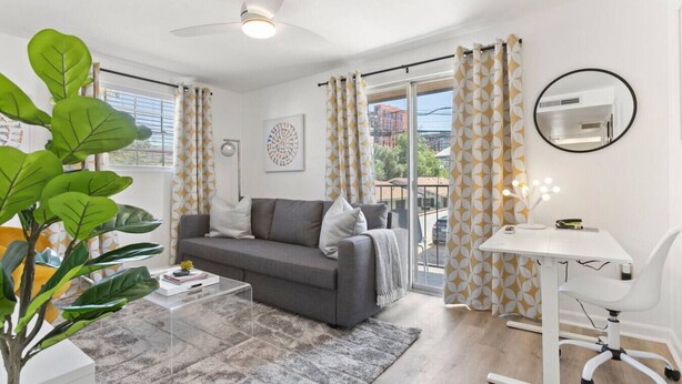 A modern living room with a grey sofa, a glass coffee table, a white desk with a mirror, and a large window with patterned curtains.