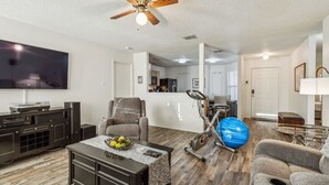 A living room with a flat-screen TV, a ceiling fan, a treadmill, and a dining area with a kitchen in the background. - Simple and Sweet 3 Bed 2 Bath (Mesquite)
