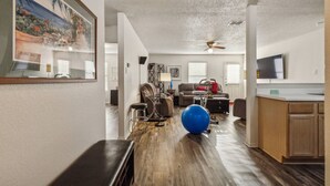 A spacious living area with a wooden floor, a ceiling fan, a large exercise ball, and a kitchen area with wooden cabinets. - Simple and Sweet 3 Bed 2 Bath (Mesquite)