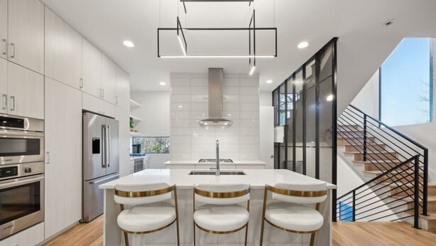 A modern kitchen with white cabinetry, stainless steel appliances, a central island with bar stools, and a staircase with a black railing.