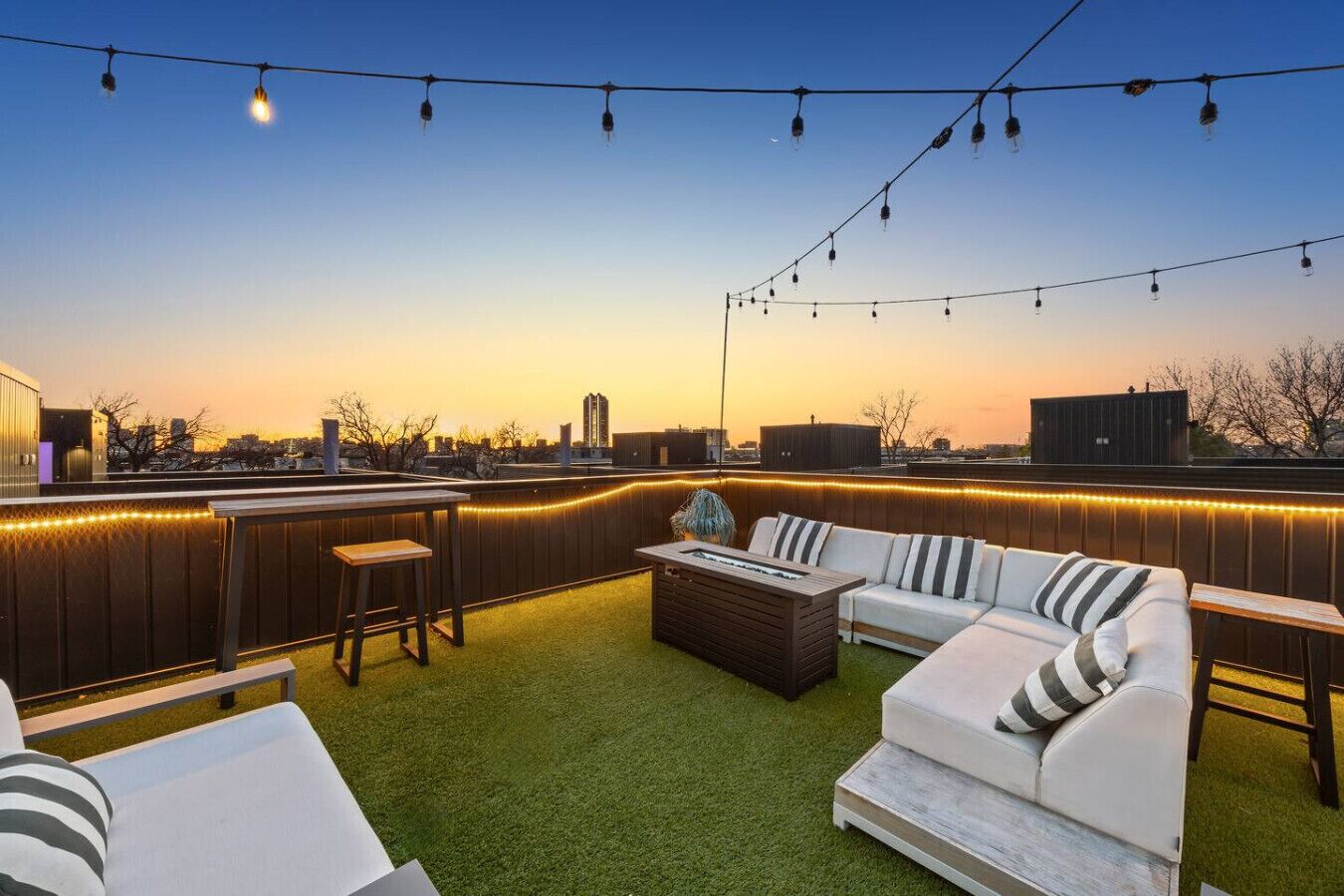 A rooftop patio with a white sofa, striped cushions, a wooden coffee table, and string lights.