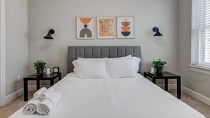 A bedroom with a large bed, two bedside tables, a grey upholstered headboard, and three framed artworks on the wall.