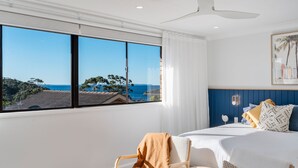A bedroom with a large window offering a view of the sea and trees, a bed with white bedding, and a ceiling fan.