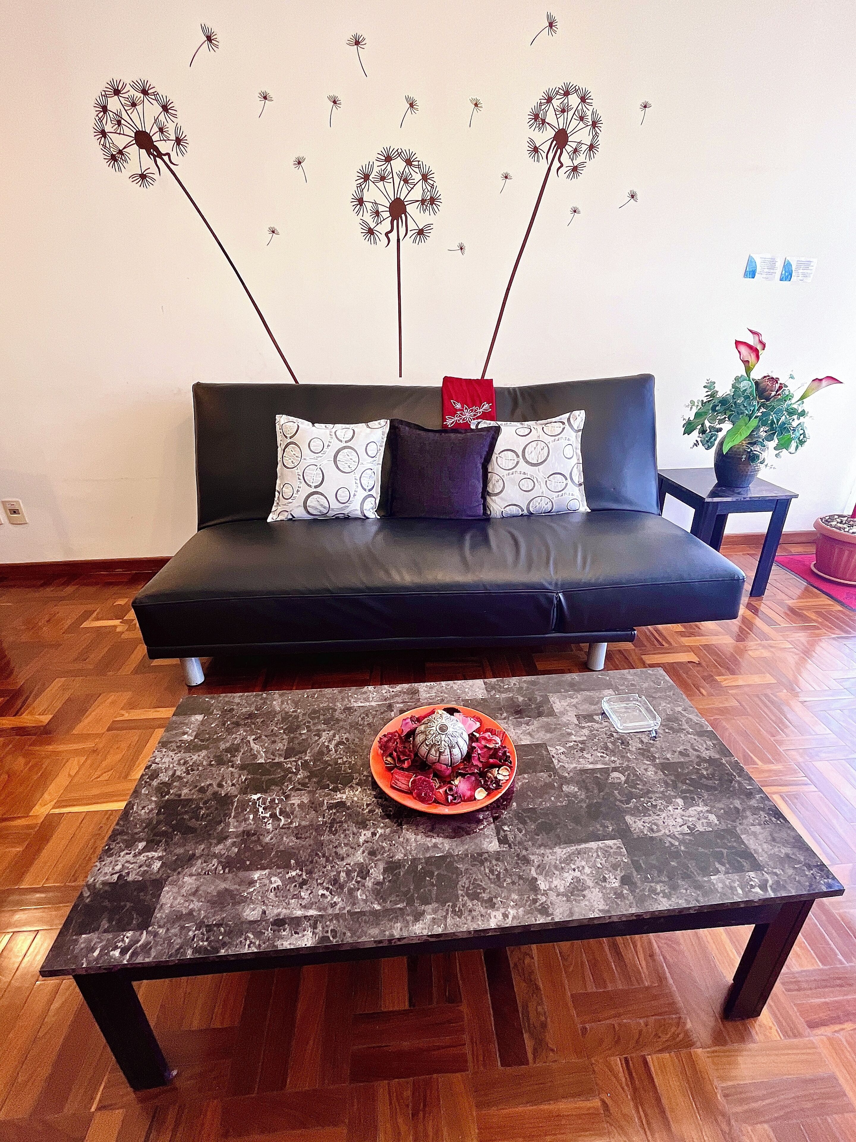 A modern living room with a dark sofa, patterned cushions, a coffee table with decorative items, and a wall with dandelion illustrations.