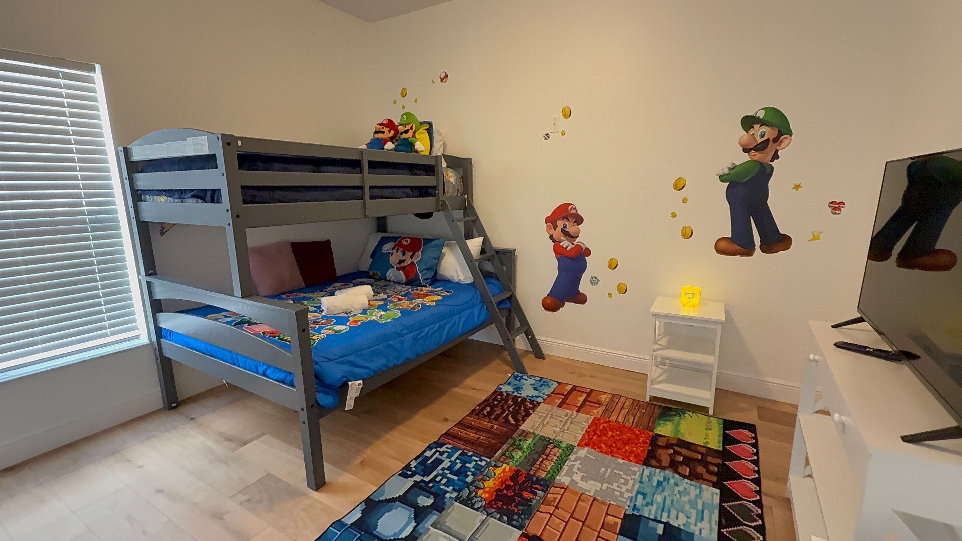 A bunk bed room with Mario and Luigi wall decals, a Minecraft-themed rug, and a TV on a white stand.