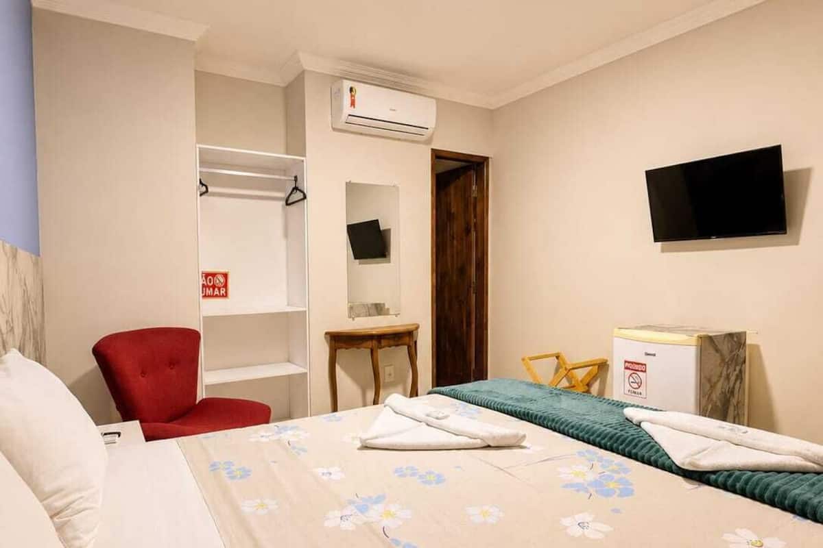 A hotel room with a bed, a red chair, a wardrobe, an air conditioning unit, a television, and a desk with a box.