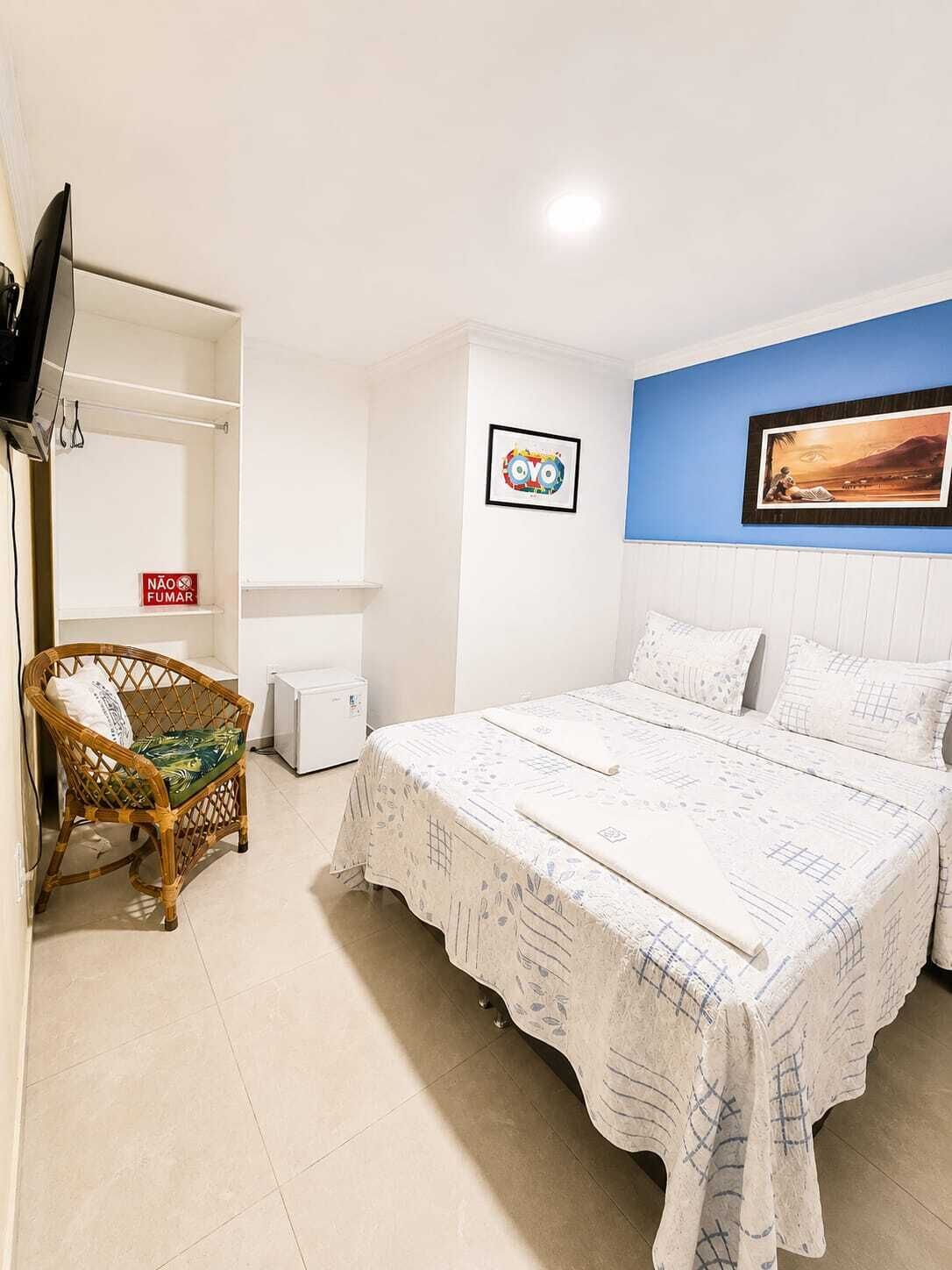 Une chambre à coucher comprenant un lit, une chaise, un téléviseur, une petite armoire blanche et un mur orné d’un tableau encadré et d’une affiche.