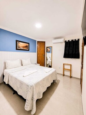A bedroom with a bed, a blue wall, a wooden door, a mirror, and a small wooden stool. - Pousada Raízes do Porto (Ipojuca)