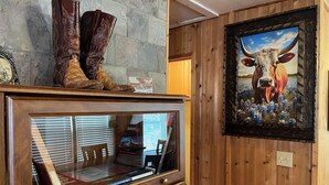 Un mur en bois orné d’un tableau encadré représentant une vache, une vitrine contenant des livres et une paire de bottes de cowboy posée sur une étagère.