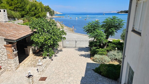 A stone-paved courtyard with a view of the sea, a gate, and surrounding greenery.