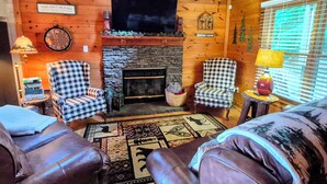A cozy cabin interior with a stone fireplace, checkered armchairs, leather sofas, and a large window with blinds. - Mountain Views! Family-friendly, 4 BR + Bunkroom, NEW Hot Tub, Firepit, Gameroom (Hiawassee)