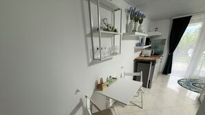 A modern kitchen with white cabinetry, a dining table, and a chair. There are shelves with decorative items and a window with curtains.