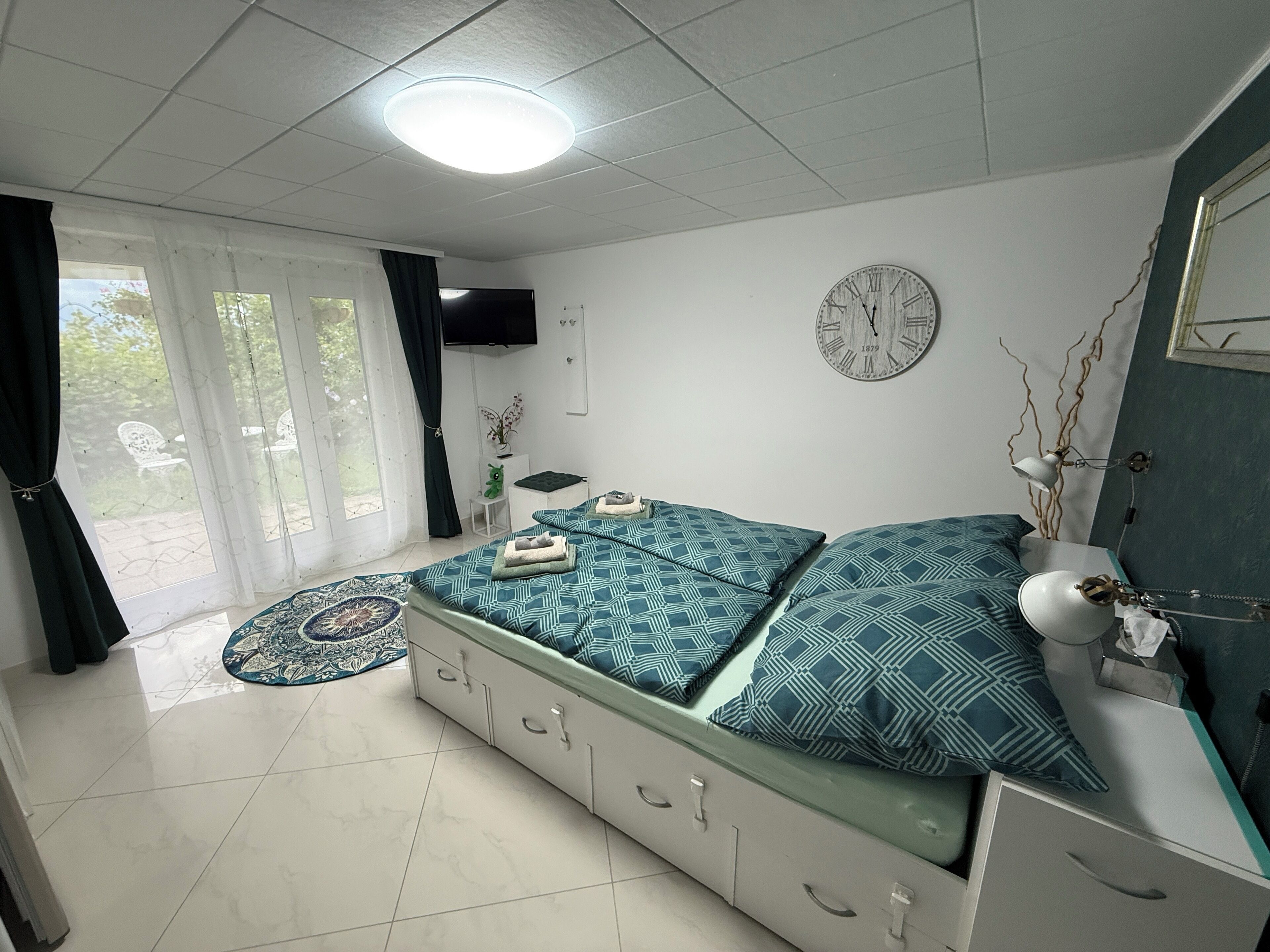 Ein Schlafzimmer mit einem Bett, einem Fernseher, einer Uhr und einem Fenster mit Vorhängen.