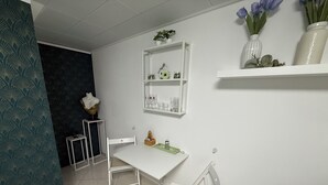 A dining area with a white table and chairs, a small shelf with decorative items, and a wall-mounted shelf with flowers.