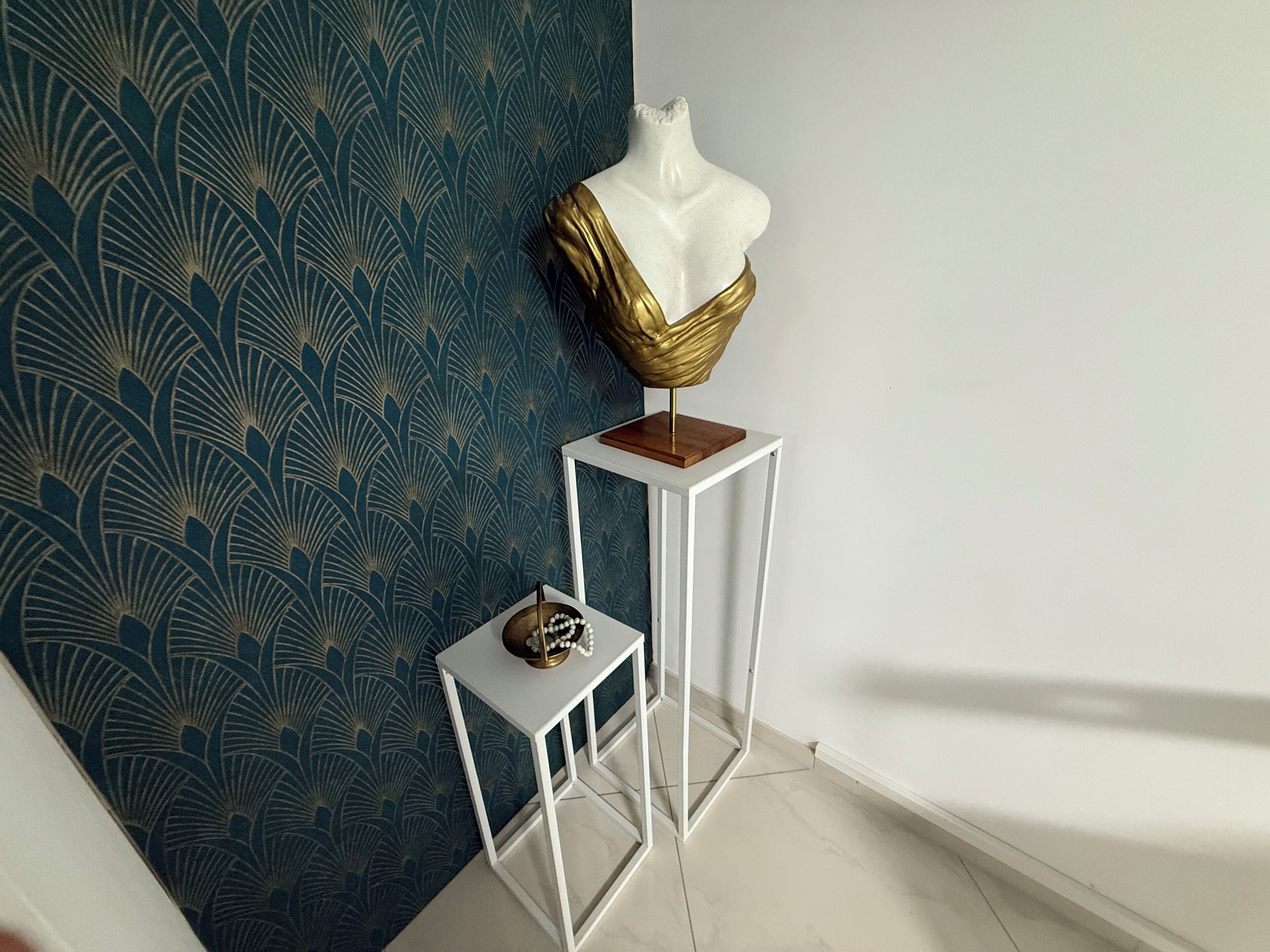 A room with a blue patterned wallpaper, a white pedestal table with a golden sculpture, and a small white table with a decorative item.