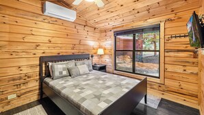 Un dormitorio de cabaña de madera con cama, mesita de noche, lámpara y ventana con vistas al exterior.