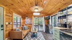 Interior acogedor de una cabaña con techo de madera, zona de estar con sofá y mesa de centro, comedor con mesa y sillas, y cocina con electrodomésticos y estantes.