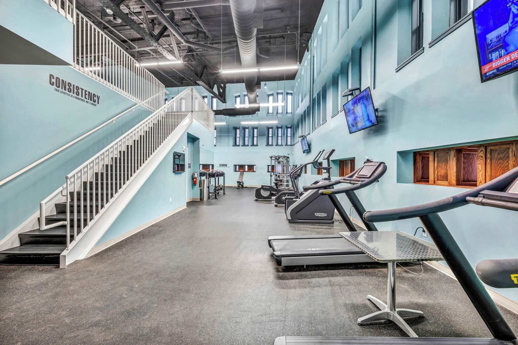 A gym with treadmills, stationary bikes, and weight machines. There is a staircase and a TV mounted on the wall.