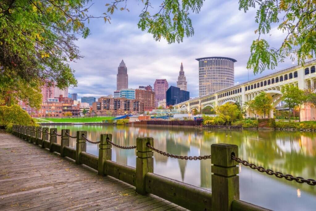 A waterfront promenade with a wooden walkway, a chain-link fence, and a city skyline in the background.